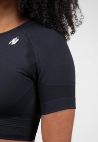 Zwarte sporttop met korte mouwen, gebreide textuur en een witte logoaccent op de schouder. Rekbaar materiaal voor comfort.