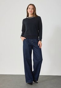 Sudadera negra de manga larga con cuello adornado, combinada con vaqueros de mezclilla azul de pierna ancha. Modelo posa con las manos en los bolsillos frente a un fondo neutro.