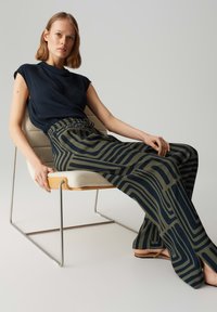 Marineblauwe blouse met een ontspannen pasvorm, gecombineerd met wijde broeken met een abstract groen-en-marineblauw patroon. Model zit op een moderne stoel.