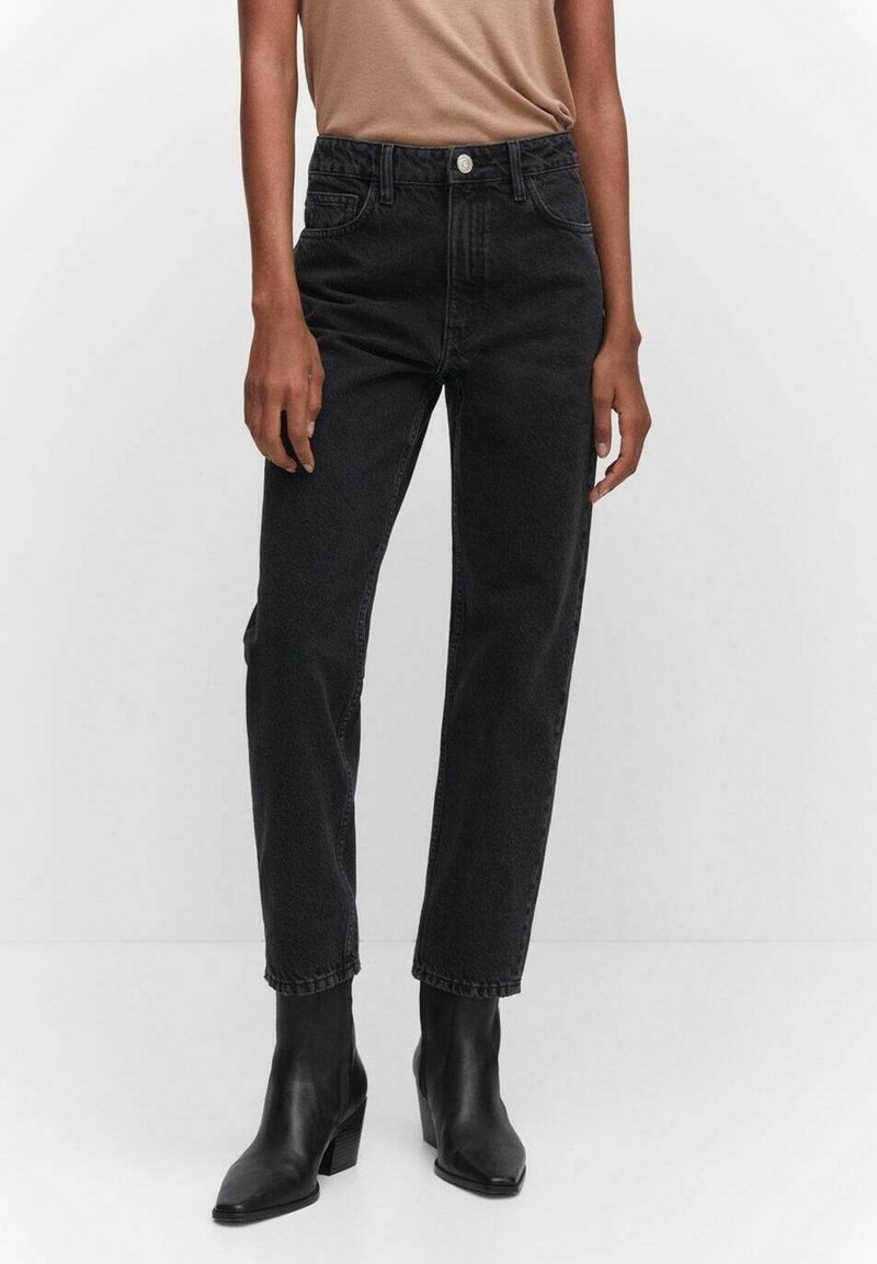 Personne portant un jean noir droit taille haute avec bouton et fermeture éclair, associé à des bottines noires à talons et un haut beige rentré.