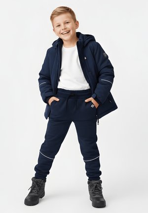 Uśmiechnięty chłopiec stoi z rękami w kieszeniach, ubrany w granatową kurtkę, pasujące spodnie z odblaskowymi detalami, białą koszulę i szare sznurowane buty.