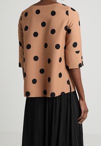 Kvinna iklädd en beige blus med stora svarta prickar och trekvartslånga ärmar, tillsammans med en svart plisserad kjol.