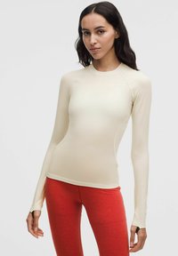 Top de manga larga color beige claro, hecho de un tejido suave y elástico. Presenta un escote redondo y mangas raglán, combinado con leggins ajustados rojos.