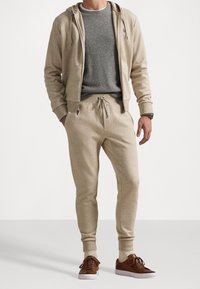 Beige hoodie med dragsko och framfickor, matchad med joggingbyxor i samma färg. Grå tröja med rund hals under. Bruna sneakers.