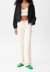 Kobieta ubrana w czarną krótką kurtkę, biały marszczony crop top, kremowe spodnie o szerokich nogawkach i jasne zielone gumowe sandały, stojąca z jedną ręką w kieszeni.