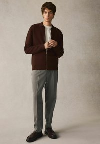 Brun zip-up striktrøje over en hvid skjorte, parret med lysegrå stribede bukser og mørkebrune loafers. Glat, blød tekstur.