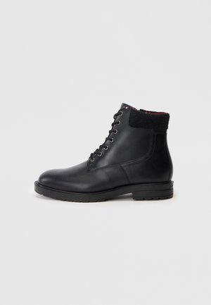 Melna ādas lace-up potenē ar mīkstinātu melnu apkakli un plakanu gumijas zoli, attēlots no ārējās puses uz baltā fona.