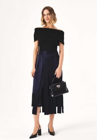 Top nero con spalle scoperte abbinato a una gonna pieghe blu navy. Borsa nera con componenti in argento. La modella indossa eleganti tacchi neri.