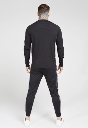 Homme debout, de dos, portant un haut à manches longues noir, un pantalon noir avec des rayures blanches sur les côtés et des baskets blanches sur un fond uni.