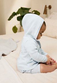 Felpa con cappuccio azzurro chiaro lavorata a maglia con una texture morbida, caratterizzata da una chiusura frontale con bottoni e polsini a coste, abbinata a pantaloncini a righe.