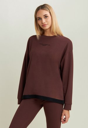 Giovane donna con capelli biondi lisci che indossa una maglia marrone a maniche lunghe e pantaloni abbinati su uno sfondo chiaro uniforme.