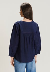 Granatowa koszula z długim rękawem wykonana z lnu, z marszczonym tyłem, koronkowymi akcentami na rękawach i elastycznymi mankietami. Noszona z jasnoniebieskimi dżinsami.