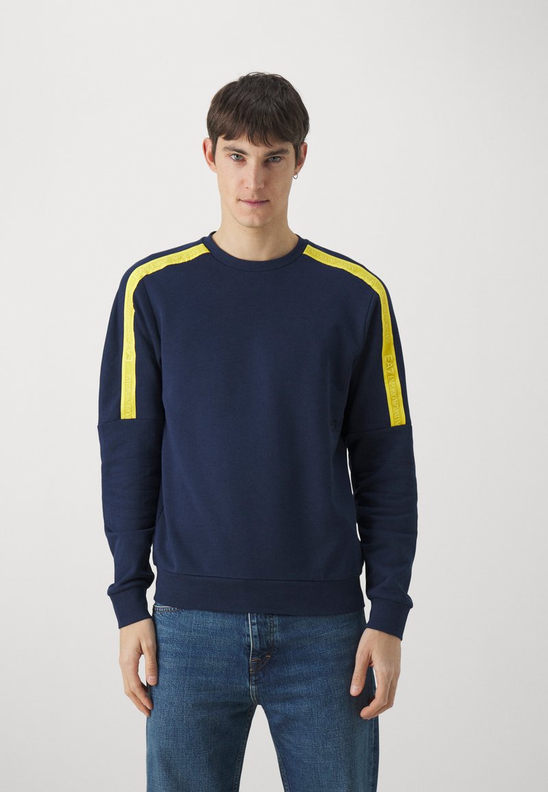 Felpa blu navy con strisce gialle laterali e tessuto texturizzato. Caratterizzata da un collo a giro, polsini a costine, abbinata a jeans blu.