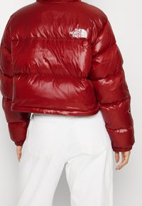 Czerwona kurtka puchowa o błyszczącej powierzchni, skróconym kroju oraz elastycznych mankietach i dolnym wykończeniu. Posiada biały logo na górnej części pleców.