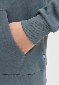 Tmavě šedá mikina s žebrovanými manžetami, postranní kapsou a malou značkovou etiketu na dolním švu, která má hladkou strukturu.