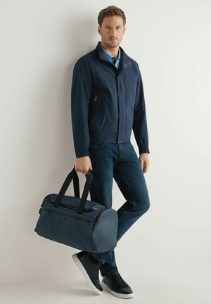 Mężczyzna w granatowej kurtce, niebieskich dżinsach i czarnych tenisówkach, trzymający granatową torbę sportową, stojący na jasnym, jednolitym tle.