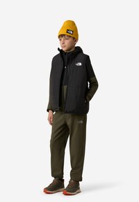 Fekete bélelt mellény fekete cipzáras dzseki fölött, olívazöld nadrág és sárga kötött sapka. Logók és kontrasztos cipőszínek jellemzik.