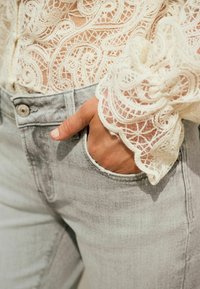 Lichtgrijze denim jeans met een voorzak, gecombineerd met een textuur blouse van ivoorkleurige kant met gedetailleerde patronen en een gegolfde mouw.