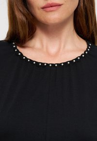 Femme portant un haut noir avec un décolleté festonné orné de petites perles blanches, montrant ses lèvres et ses clavicules.