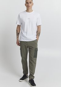 White cotton t-shirt with small reflective text, paired with olive green cargo pants featuring side pockets and black logo, complemented by black sneakers.