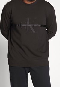 Czarna bluza z haftowanym logo "CK" oraz napisem "CALVIN KLEIN JEANS" na satynowej taśmie. Prążkowane mankiety i dół, miękki materiał.