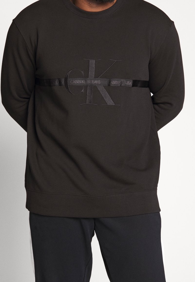 Czarna bluza z haftowanym logo "CK" oraz napisem "CALVIN KLEIN JEANS" na satynowej taśmie. Prążkowane mankiety i dół, miękki materiał.