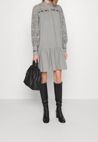 Vestido gris con falda acampanada y mangas largas con patrón de ojales, combinado con botas negras hasta la rodilla y un bolso de cuero negro.
