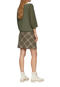 Haut vert olive avec des manches texturées, associé à une jupe à carreaux aux tons verts et crème, et des bottines lacées blanches.