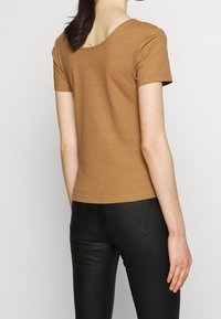 Ribbede brune toppe med rund halsudskæring, korte ærmer, tætsiddende design. Bæres med sorte jeans med baglommer og syningsdetaljer.