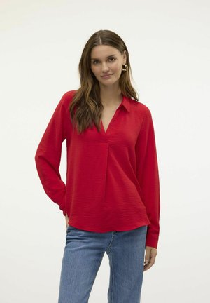 Blusa rossa a maniche lunghe con colletto, tessuto texturizzato e scollatura a V. Abbinata a jeans blu per un contrasto.