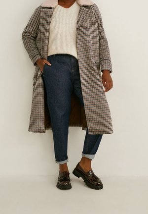 Personne portant un pull beige, un manteau long à carreaux avec un col en peau retournée, un jean bleu foncé retroussé et des mocassins marron à glands, debout contre un mur blanc.