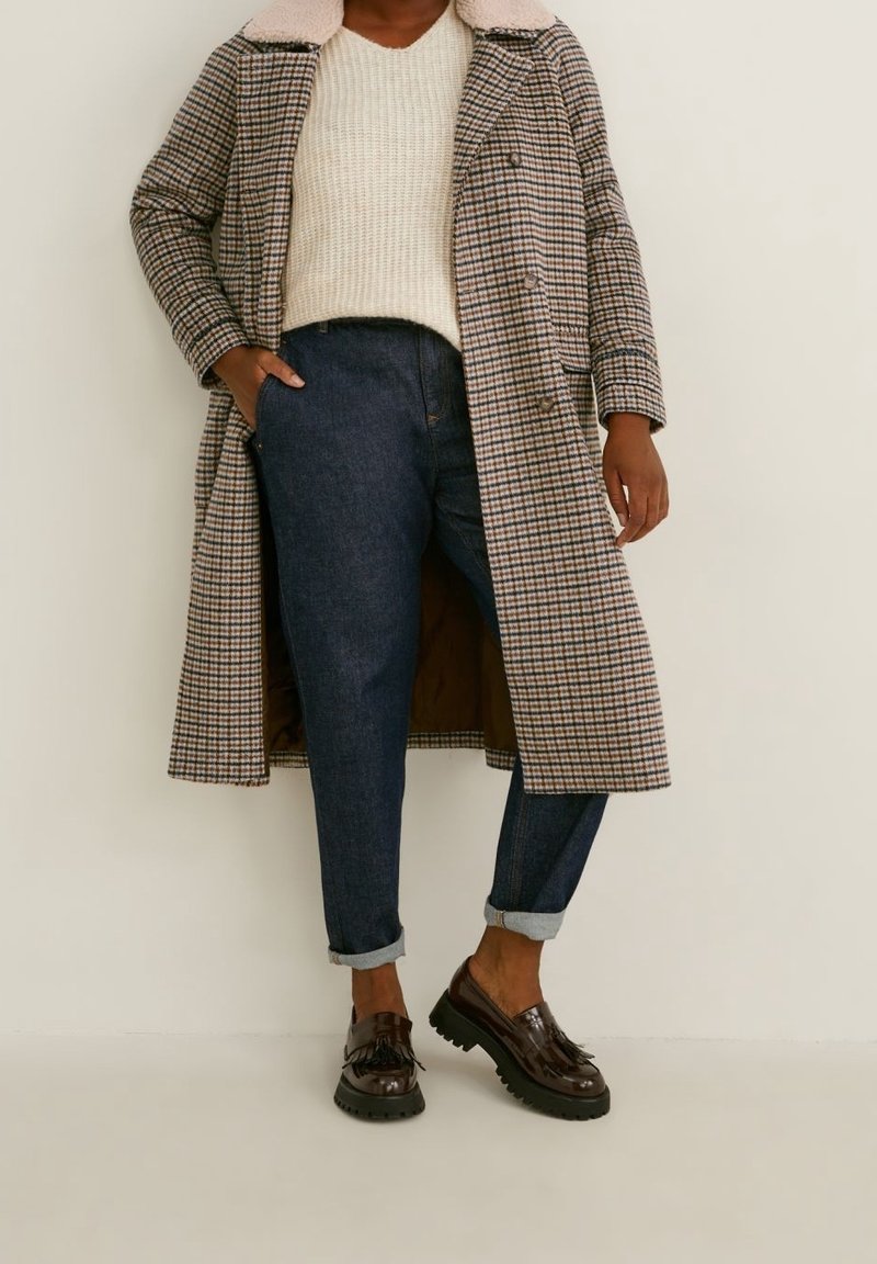 Personne portant un pull beige, un manteau long à carreaux avec un col en peau retournée, un jean bleu foncé retroussé et des mocassins marron à glands, debout contre un mur blanc.