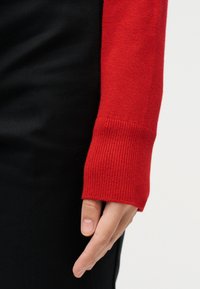 Roka sproščeno ob telesu, oblečena v rdeč pleteni rokav puloverja in črne hlače na belem ozadju.