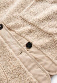 Tessuto in pile beige con superficie texturizzata, dotato di una fodera liscia color marrone chiaro e due bottoni neri per la chiusura. Una tasca visibile.