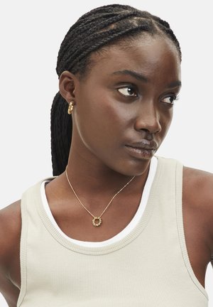 Goldene Creolen und eine zarte goldene Halskette mit einem runden Anhänger. Der Betreff trägt ein beigefarbenes geripptes Tanktop über einem weißen Hemd.