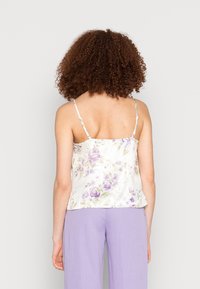 Femme aux cheveux bouclés portant un débardeur floral blanc et un pantalon lavande, se tenant de dos devant un fond clair uni.