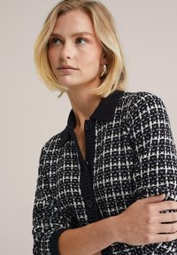 Pletený sveter s čiernobielym kockovaným vzorom, rebrovaným golierom a manžetami, predným zapínaním na gombíky; dlhé rukávy s voľným strihom.