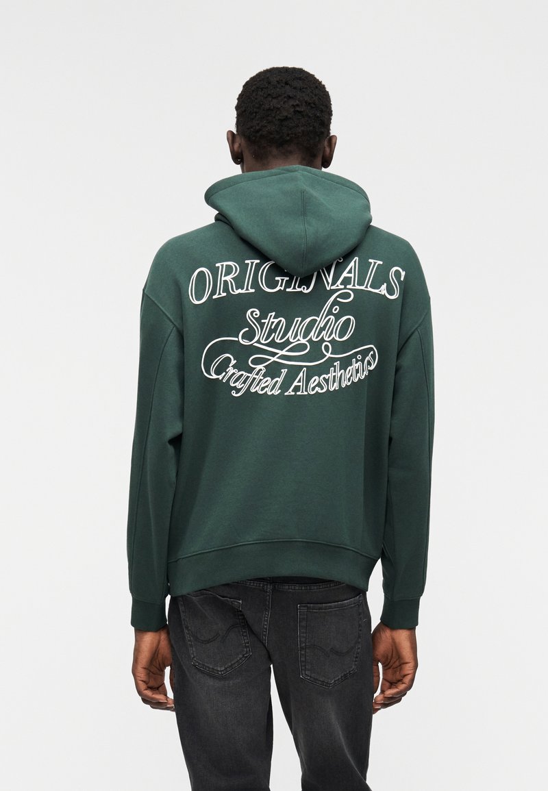 Το πράσινο φούτερ με κουκούλα διαθέτει κουκούλα, υπερμεγέθη εφαρμογή και λευκό κείμενο στην πλάτη που γράφει "ORIGINALS Studio Crafted Aesthetics." Υλικό από βαμβακερό μείγμα.