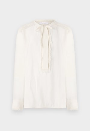 Blusa bianca con colletto rotondo, dettaglio collo annodato e maniche lunghe. Presenta accenti di frange testurizzate lungo le spalle e le maniche.