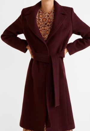 Femme portant un manteau ceinturé bordeaux foncé sur une robe imprimée serpent marron, mains posées sur les hanches, debout devant un fond uni.