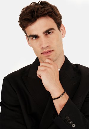 Blazer nero con rever affilati, indossato su una camicia bianca. Modello maschile posa con una mano sul mento, mostrando un braccialetto di perle nere sul polso.
