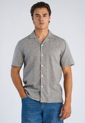 Mand iført en kortærmet, lysegrå skjorte med knapper, stoppet ned i blå jeans, med den ene hånd i lommen mod en ensfarvet baggrund.