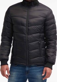 Giubbotto nero imbottito con colletto alto e chiusura a zip. Presenta un design trapuntato orizzontale e polsini a coste. Indossato sopra jeans blu.