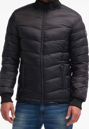 Giubbotto nero imbottito con colletto alto e chiusura a zip. Presenta un design trapuntato orizzontale e polsini a coste. Indossato sopra jeans blu.
