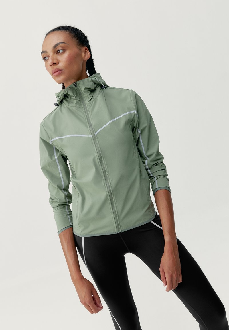 Svetlozelená dlhá bunda s dlhým rukávom vyrobená z vodoodolného materiálu, s zipsom, kapucňou a reflexnými bielymi detailmi. V kombinácii s čiernymi legínami.