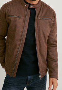 Mann trägt eine braune Lederjacke mit Reißverschlusstaschen über einem schwarzen, strukturierten Pullover und blauer Jeans, steht mit einer sichtbaren Hand.