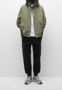 Olijfgroene windjack met een hoge kraag en zijzakken, gecombineerd met zwarte cargobroek. Grijze sneakers met witte accenten en veters.