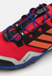 Rote Sportschuh mit strukturiertem Obermaterial, blauen und schwarzen Schnürsenkeln sowie orangefarbenen Akzenten. Enthält GORE-TEX-Branding und eine strapazierfähige Sohle.