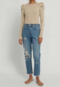 Beige gerippter Langarmtop mit Puffärmeln, kombiniert mit stark ausgewaschenen hellblauen Jeans, die fransige Säume und mehrere Risse aufweisen.