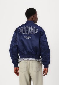 Chaqueta bomber satinada en color navy con puños y dobladillo elásticos. "ORIGINALS" bordado en la espalda, con cuello tipo crew, superpuesta sobre una camisa de cuadros.
